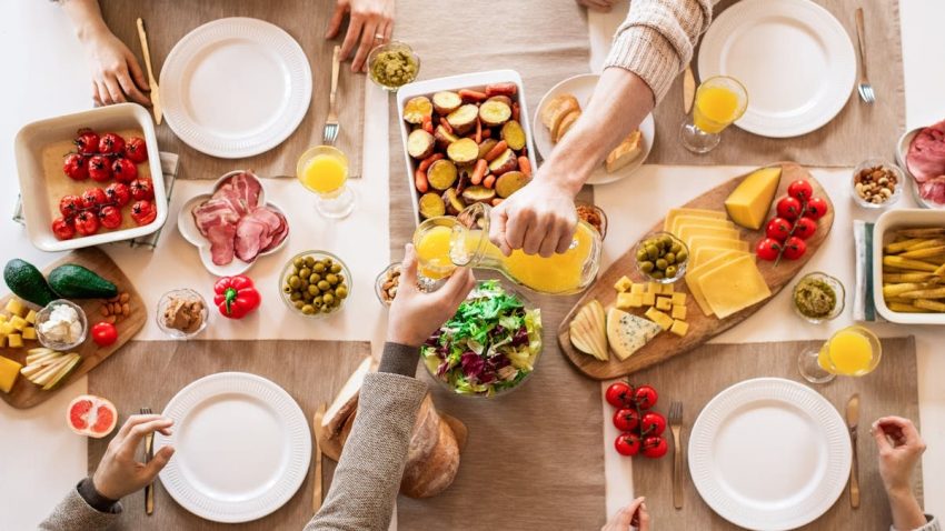 Overhead view of a lively breakfast table featuring orange juice, cheeses, fresh vegetables, and cured meats.